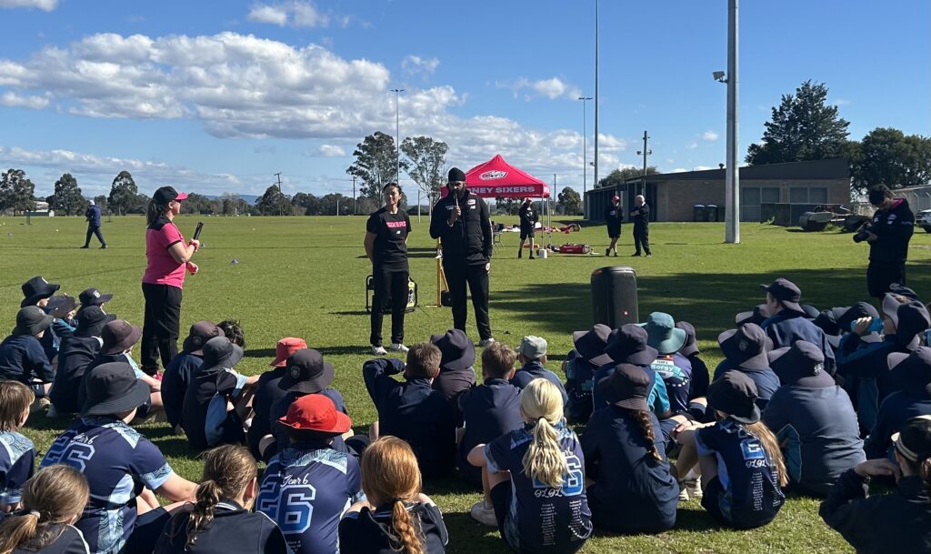 Students from Catholic schools sit on a sports field listening to Sydney Sixers representatives during a Schools Smash Cricket Gala Day activity.